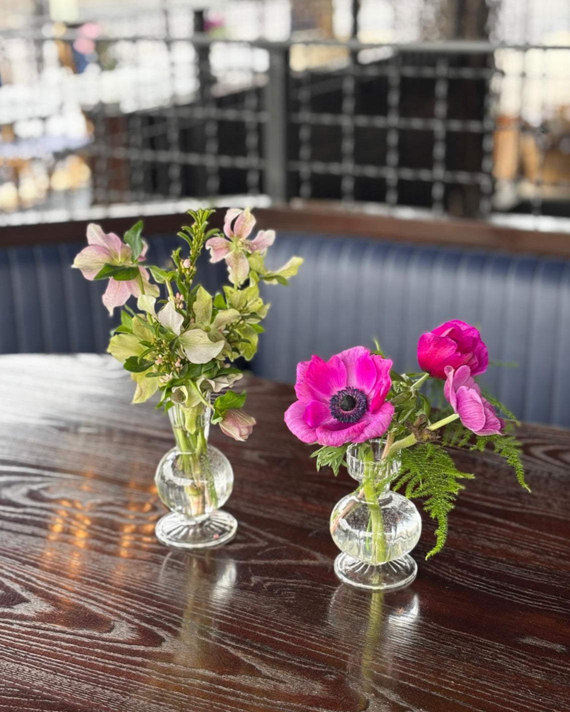 Two small glass bud vases with flowers on a wooden table, blurred restaurant interior in the background.