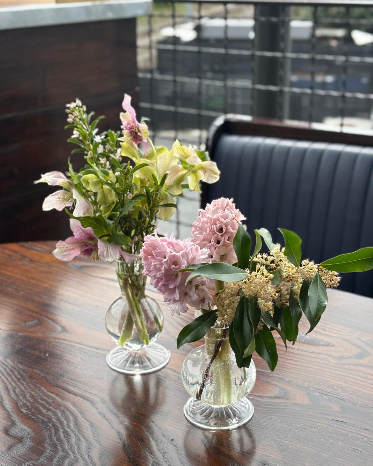 Two flower arrangements in glass vases on a wooden table with a blurred outdoor background.