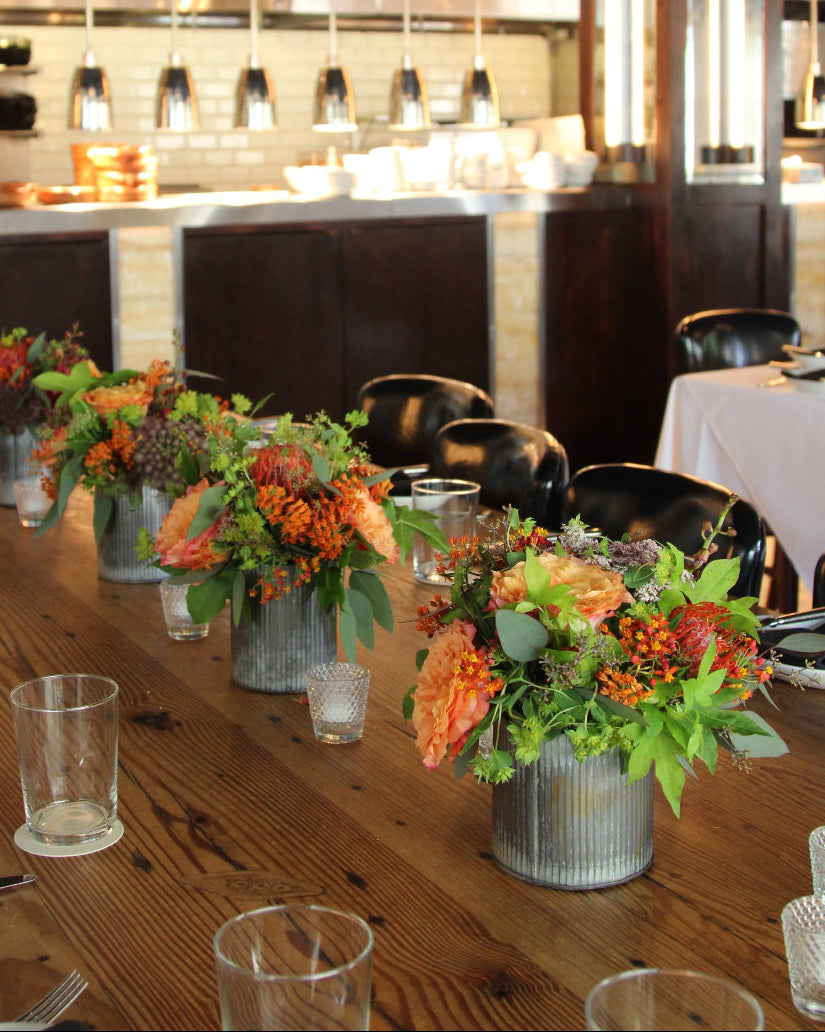 Top-down view of a wooden table with floral centerpieces and glasses, reflecting a restaurant or bar setting.