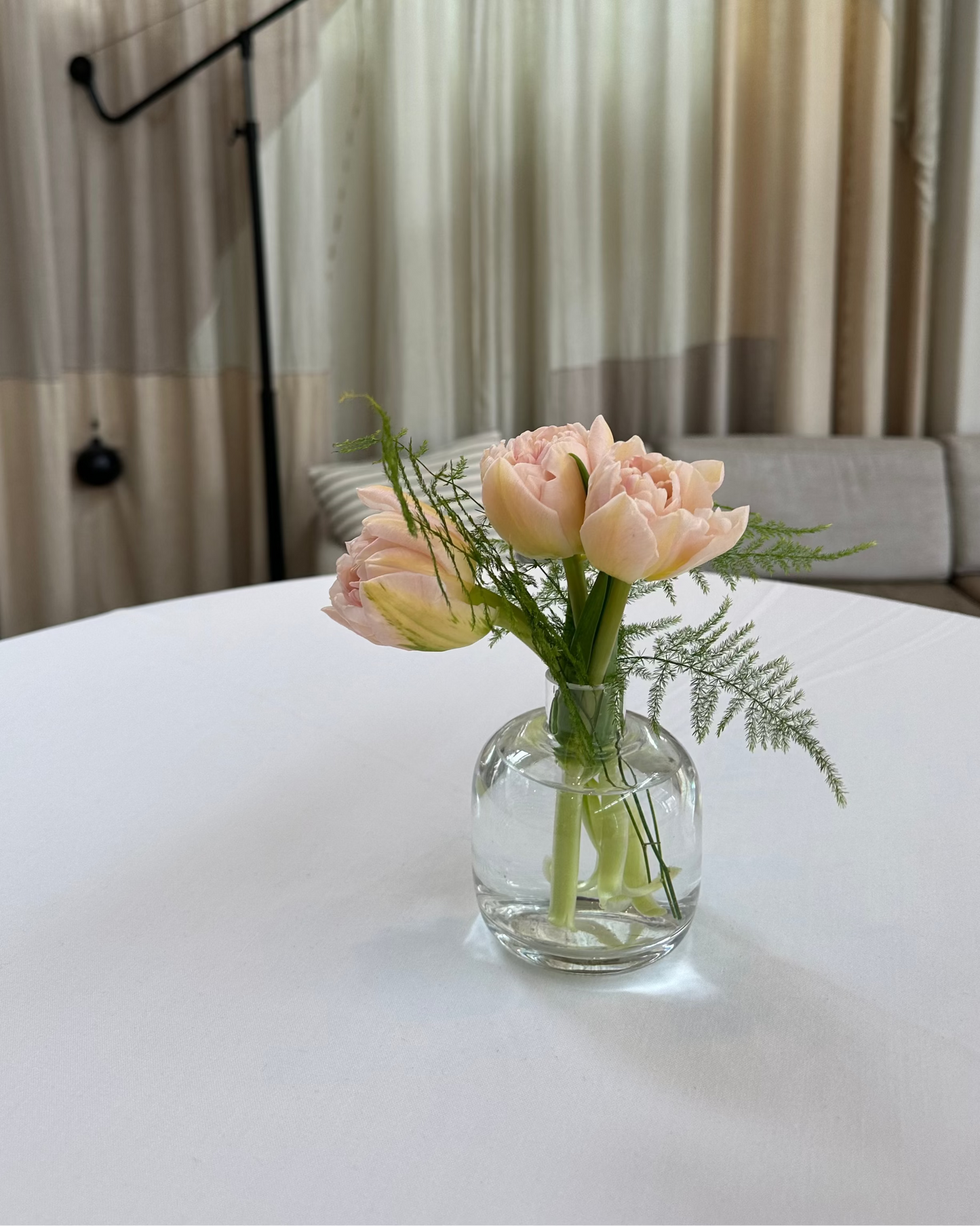 Bouquet of pink flowers in a clear vase on a white table with a lamp and curtains in the background.